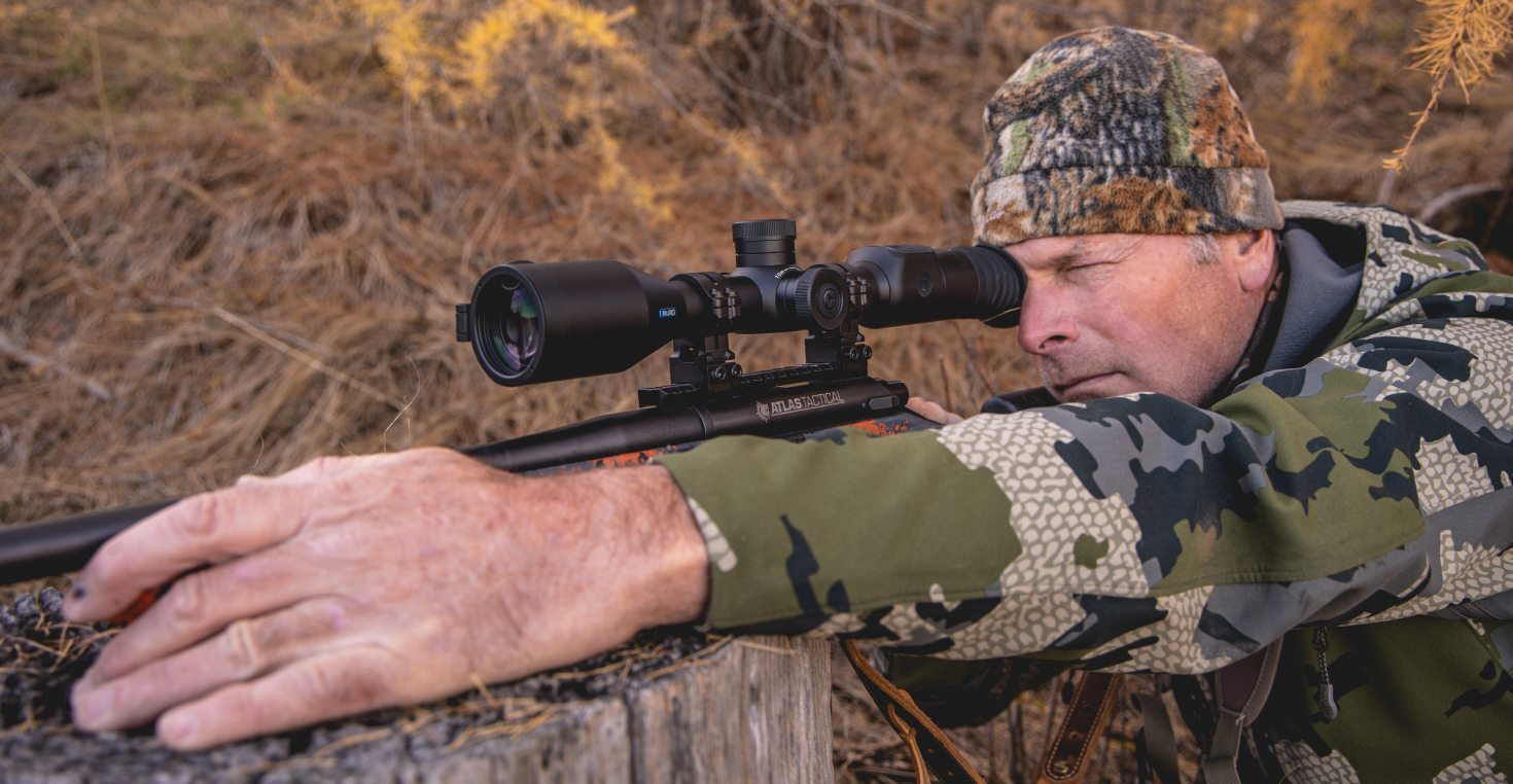 Hunter Zeroing In with the PARD Nightstalker EX Scope A hunter in camouflage gear lines up a shot using a PARD Nightstalker EX mounted on a rifle, positioned over a tree stump in a dry grass field. The image highlights real-world use, stable shooting posture, and the clarity-focused design of the night vision scope.
